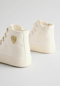 Cremefarbene High-Top-Sneaker aus Canvas mit einem goldenen Herz-Patch, Schnürsenkeln und einem seitlichen Reißverschluss für einfachen Zugang.