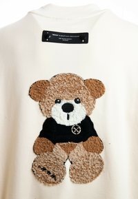 Sudadera color crema con un aplique de un oso de peluche marrón texturizado que lleva una camiseta negra con un logotipo circular.
