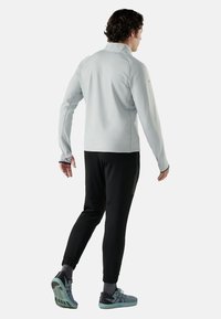 Lichtgrijs langetermijn shirt met hoge kraag, gecombineerd met strakke zwarte broeken. Heeft elastische manchetten en lichtblauwe sportschoenen met donkere accenten.
