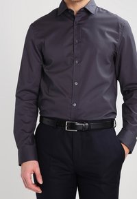 Camicia a maniche lunghe con bottoni di colore grigio scuro, dal tessuto liscio, con colletto a punta e bottoni neri. Indossata con pantaloni blu navy e una cintura nera.