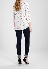 White blouse with bird pattern, three-quarter sleeves, ruffled neckline. Worn with dark skinny jeans and black high-heeled sandals.