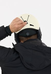 Hvid hjelm med sorte detaljer og ventilationsåbninger, justeres af en hånd og bæres over en sort jakke med hætte.