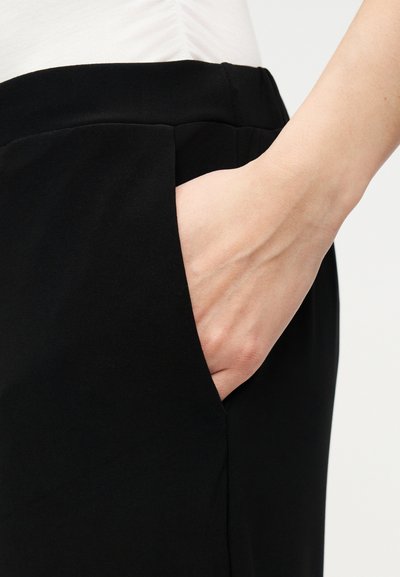 Χέρι που ακουμπά στην δεξιά τσέπη μαύρου παντελονιού που φοριέται με λευκό τοπ, απεικονιζόμενο από τη μέση και κάτω.