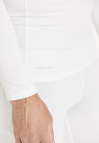 Haut de sport blanc à manches longues avec une étiquette "Sèche Rapide". Le matériau est lisse avec une coupe ajustée et une texture subtile près des coutures.