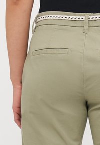 Calças verde claro com um bolso traseiro, acompanhadas por um cinto texturizado com um design trançado em preto e branco.