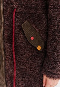 Hnědá fleecová bunda s texturou, hnědým manžetovým víčkem na kapse z kordu upevněným červeným knoflíkem, červeným pleteným zipem a rukou osoby odpočívající na boku.