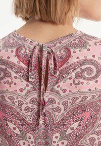 Roze paisleyblouse met een ronde halslijn, een stoffen strik op de rug en complexe bloemmotieven. Zachte textuur en vloeiende lijn.