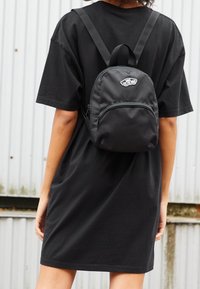 Osoba oblečená v černé přehnaně velké tričkové šaty a s malým černým batohem Vans stojí u vlnité kovové zdi.