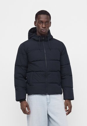 Homme portant une veste matelassée noire à capuche avec une fermeture éclair à l’avant et un jean bleu clair, debout devant un fond blanc uni.