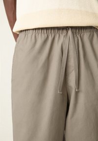 Lichtbruine, lichte broek met een elastische tailleband en koordje. Heeft een gladde textuur en geen zichtbare patronen of accenten.