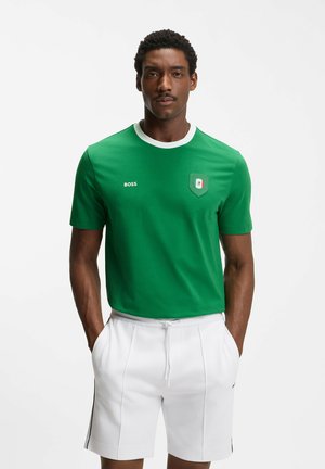 Mann trägt ein grünes BOSS-T-Shirt mit Wappen und weiße Shorts, steht mit den Händen in den Taschen vor einem schlichten weißen Hintergrund.