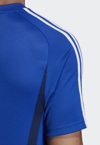 Blaues Sportshirt mit glatter Textur, kurzen Ärmeln und weißen Streifen an den Schultern. Dunkle Seitenpaneeldetails sichtbar.