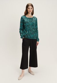 Mujer de pie vestida con una blusa de manga larga en teal con estampado paisley, pantalones negros cropped de pierna ancha y bailarinas beige, sobre un fondo neutro.