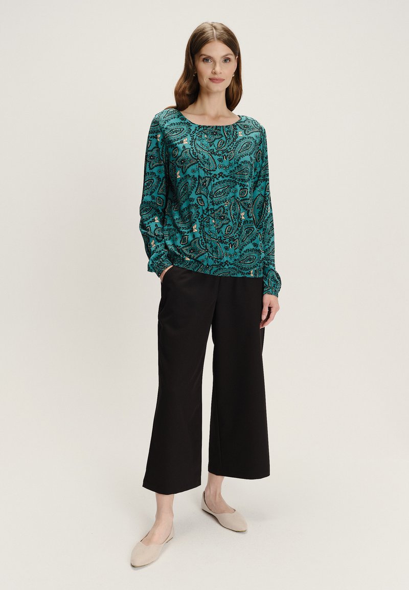 Mujer de pie vestida con una blusa de manga larga en teal con estampado paisley, pantalones negros cropped de pierna ancha y bailarinas beige, sobre un fondo neutro.