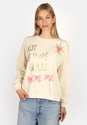 Woman with blonde hair wearing a cream sweatshirt with gold and red text and a star with heart design, paired with light blue jeans.