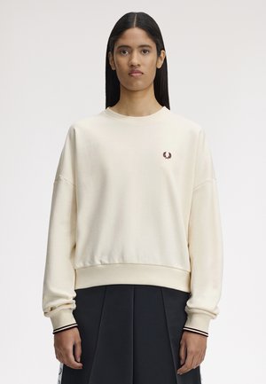 Jeune personne aux cheveux longs portant un sweat-shirt crème à col rond avec un petit logo de laurier bordeaux sur la poitrine et une jupe plissée noire.