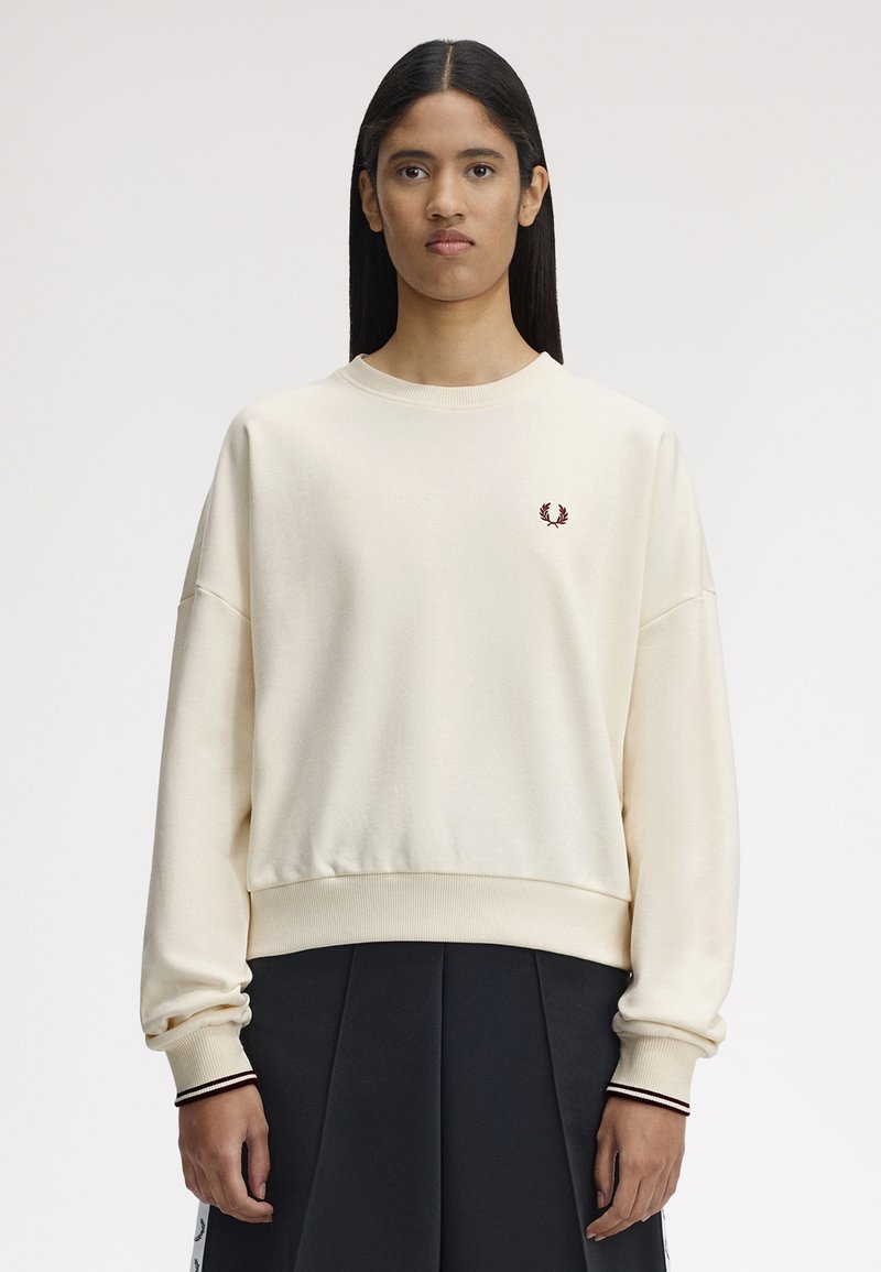 Jeune personne aux cheveux longs portant un sweat-shirt crème à col rond avec un petit logo de laurier bordeaux sur la poitrine et une jupe plissée noire.