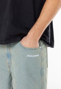 Light blue denim pants with a fitted shape, featuring a front pocket and white "PEGADOR" text logo near the pocket seam.