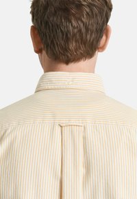 Camicia a righe giallo chiaro e bianco vista da dietro. Presenta un colletto con bottoni e una piega centrale. Tessuto dalla texture morbida.