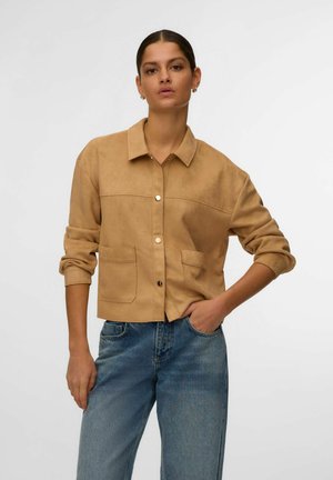 Kobieta z ciemnymi włosami związanymi do tyłu, ubrana w beżową kurtkę na guziki z podwiniętymi rękawami i niebieskie dżinsy, stojąca na tle jasnego, jednolitego tła.