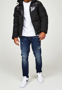 Svart puffjacka med glansiga frampaneler, vit t-shirt, blå slitna jeans och vita sneakers med svarta accenter.