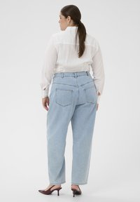 Frau mit braunen Haaren im Pferdeschwanz, die eine durchsichtige weiße langärmlige Bluse, hellblaue hoch taillierte Jeans und burgunderrote Kitten-Heels trägt, steht mit dem Rücken zur Kamera.