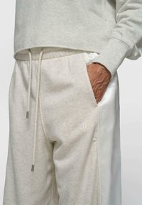 Person trägt hellgraue Jogginghose mit Kordelzug und Hand in der Tasche, mit einer kleinen "10" nahe der Tasche gestickt.