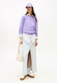 Felpa viola, camicia bianca con colletto, gonna lunga in denim chiaro con spacco frontale, sneakers bianche con suola in gomma e borsa beige.