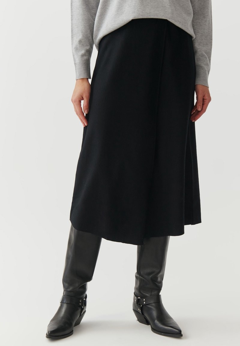 Personne portant une jupe midi noire, un haut gris à manches longues et des bottes en cuir noires montant jusqu'aux genoux, debout sur un fond clair uni.