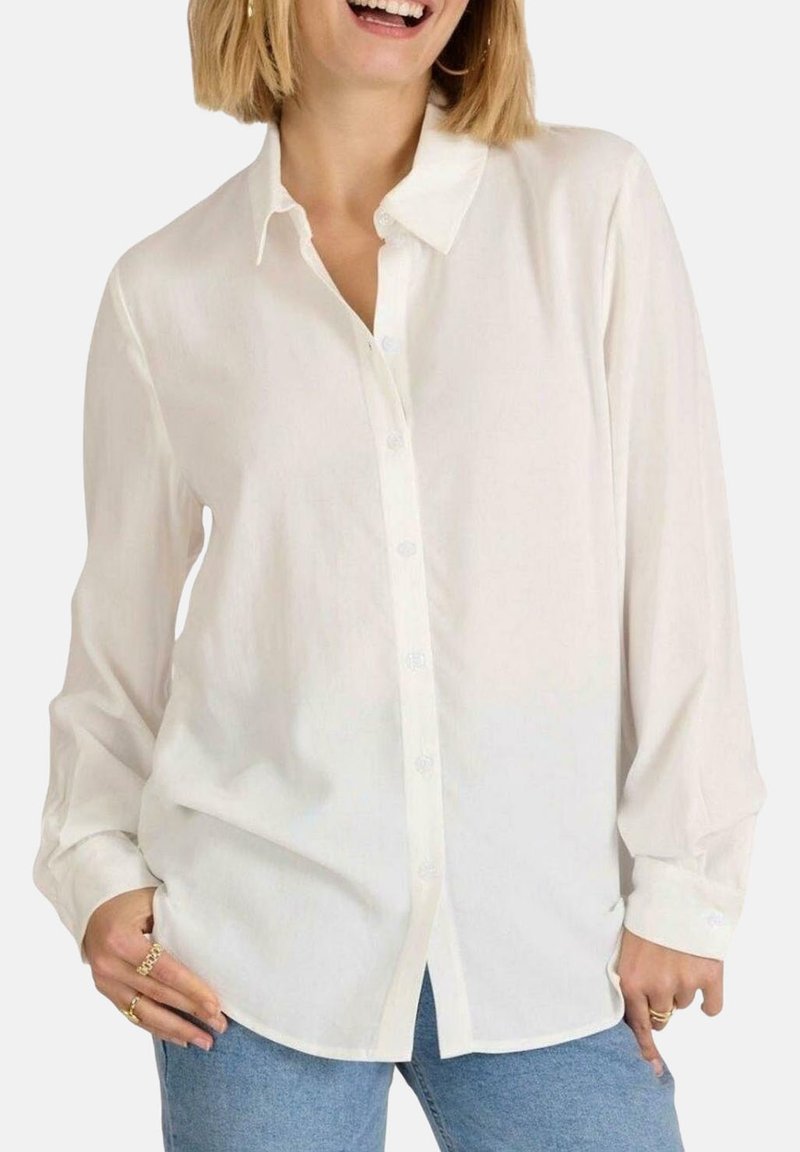 Camisa blanca de botones hecha de tela ligera, con un collar clásico, mangas largas y un corte relajado, combinada con jeans azules.
