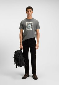Harmaa T-paita, jossa on susi-grafiikka ja kehysdesign, mustat corduroypö Pants, musta reppu, ruskeat nauhakenkiä, yksinkertainen tausta.