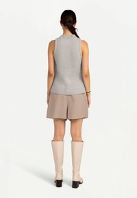 Femme aux cheveux foncés attachés en queue de cheval, portant un haut en maille gris sans manches, un short beige et des bottes crème montantes jusqu'aux genoux, debout de dos sur fond blanc.