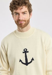Pull crème avec un col côtelé, présentant un design d'ancre bleu foncé bien en vue à l'avant. Tissu tricoté texturé.