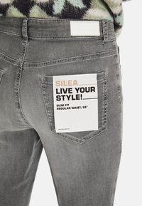 Grijze slim-fit jeans met een achterzaklabel. Label zegt "SILEA LIVE YOUR STYLE! REGULAR WAIST/28". Gestructureerde denimstof is zichtbaar.