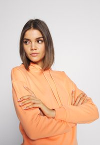 Oranžová mikina s kapucňou a šnúrkou, mäkká textúra a rebrované manžety. Modelka pózuje so založenými rukami na svetlo sivej pozadí.