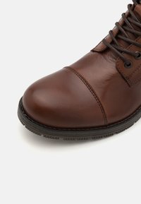 Bottines en cuir marron avec un bout arrondi, des accents cousus et des lacets foncés. La semelle est texturée avec un léger talon pour l'adhérence.