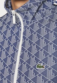 Detailní záběr na modro-bílou bundu s geometrickým vzorem, bílým zipem a malým zeleným krokodýlem na hrudi.