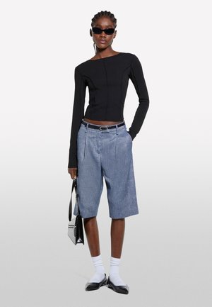 Top nero a maniche lunghe con un design attillato, abbinato a pantaloni corti plissettati in chambray blu. Gli accessori includono una borsa nera e occhiali da sole.