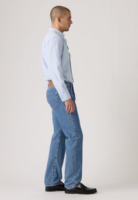 Ανοιχτό γαλάζιο πουκάμισο με κουμπιά, μπλε τζιν παντελόνι με ίσια γραμμή και μαύρα παπούτσια. Το μοντέλο είναι τοποθετημένο πλάγια, παρουσιάζοντας τη γραμμή και τα υλικά των ρούχων.