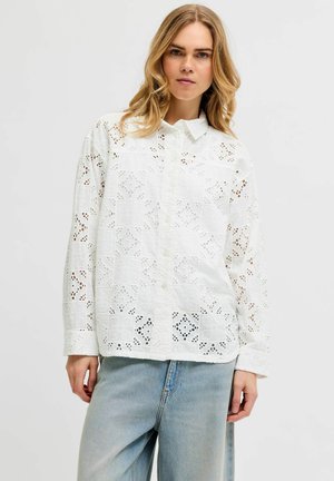 Mujer con camisa blanca holgada de encaje ojete abotonada y vaqueros azul claro, de pie contra un fondo liso y claro.