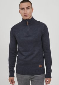 Marineblauer Strickpullover mit hohem Kragen, halber Reißverschluss vorne und gerippten Bündchen sowie Saum. Verfügt über ein braunes Logo-Patch an der Seite.