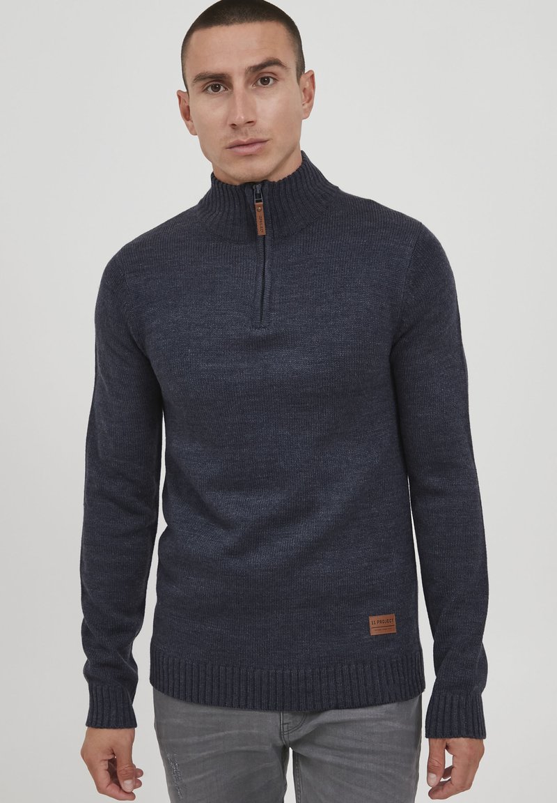 Marineblauer Strickpullover mit hohem Kragen, halber Reißverschluss vorne und gerippten Bündchen sowie Saum. Verfügt über ein braunes Logo-Patch an der Seite.
