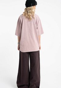 Blondynka w czarnej czapce beanie, oversize'owej jasnoróżowej koszulce i szerokich ciemnobrązowych spodniach, stojąca tyłem na białym tle.