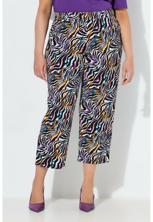 Frau trägt verkürzte Hosen mit bunten Zebrastripes in Schwarz, Weiß, Orange, Lila und Blau, kombiniert mit lila High Heels.