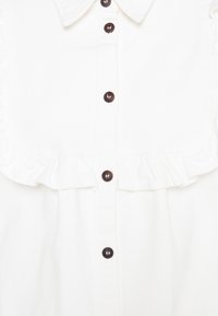 Hvid bomuldsbluse med flæsekrave og knapstolpe, lukkes med mørkebrune knapper for kontrast. Glat tekstur og struktureret design.