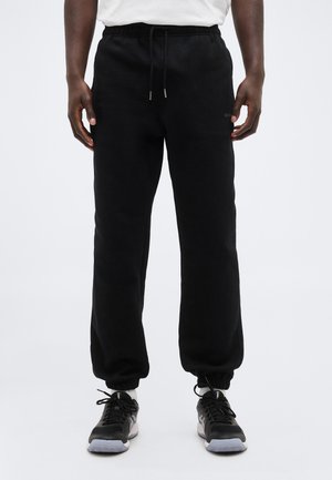 Dolna połowa osoby ubranej w czarne joggery z elastycznymi mankietami oraz czarne buty sportowe z białymi podeszwami.