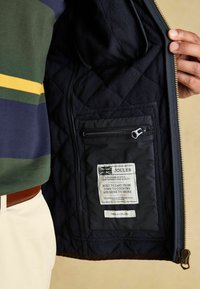 Čierna prešívaná fleecová bunda s podšívkou a bočným zipsovým vreckom. Na štítku je značka "Joules" s textom, dizajnom britskej vlajky a zipsovým uzáverom.
