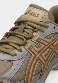 Κοντινό πλάνο ταμπά και μπεζ αθλητικού παπουτσιού Asics με λεπτομερή ραφές, στρωματοποιημένα υλικά και δέσιμο με κορδόνια.