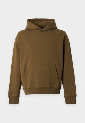 Homme portant un sweat à capuche marron et un pantalon large marron foncé, debout devant un fond blanc uni avec une main dans la poche.