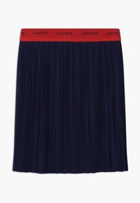 Námořnická plisovaná sukně s červeným elastickým pasem, na kterém je logo "Lacoste". Tkanina má hladkou texturu a vertikální sklady.
